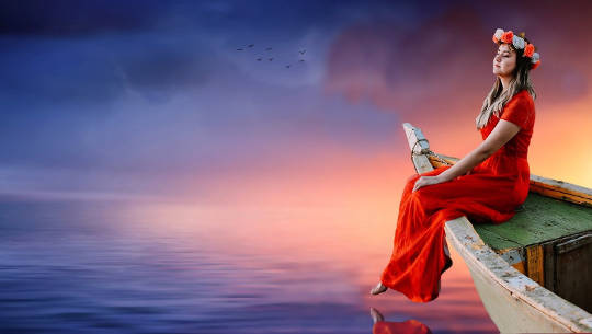 woman wearing a long dress and a garland of flowers on her head is sitting on the edge of a floating rowboat at sunset