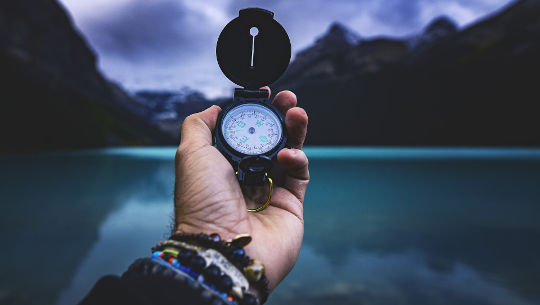 a hand holding a compass with a lake and mountains in the background
