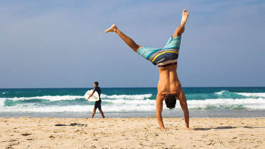 two people on a beach: one carrying a surfboard, the other doing a cartwheel