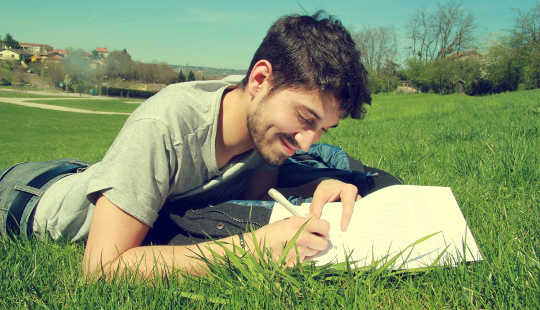 smiling man laying on the grass writing in a notebook